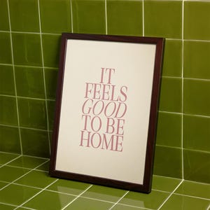 May include: A framed print with the text "IT FEELS GOOD TO BE HOME" in a soft pink hue. The frame is dark brown, and the print is leaning against a green tiled wall.