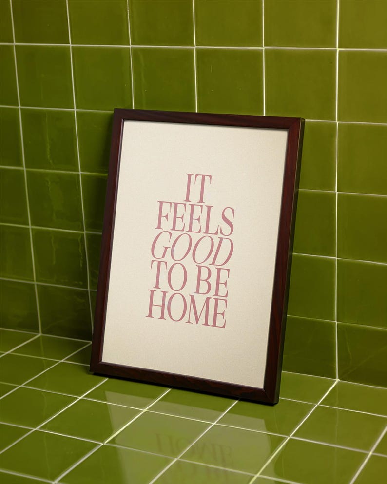 May include: A framed print with the text "IT FEELS GOOD TO BE HOME" in a soft pink hue. The frame is dark brown, and the print is leaning against a green tiled wall.