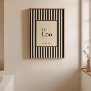 May include: A framed wall art piece with a beige background and the words "The Loo at No. 22" in black. The frame has a striped black and white pattern with a natural wood border. The art is hanging on a beige wall.