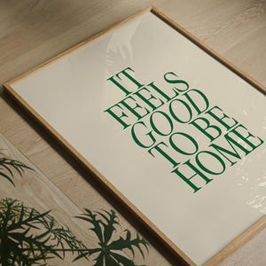 May include: A framed print with the text "IT FEELS GOOD TO BE HOME" in green lettering on an off-white background. The frame is made of light-colored wood. The image is on a wooden floor.