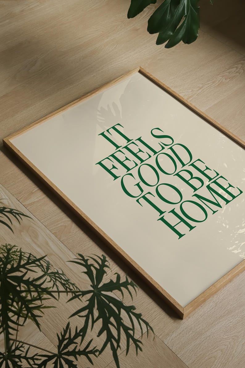 May include: A framed print with the text "IT FEELS GOOD TO BE HOME" in green lettering on an off-white background. The frame is made of light-colored wood. The image is on a wooden floor.