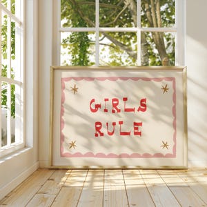 May include: A framed print with the words "GIRLS RULE" in red, stylized lettering. The print has a light pink scalloped border and gold star accents on a cream background. The frame is gold.