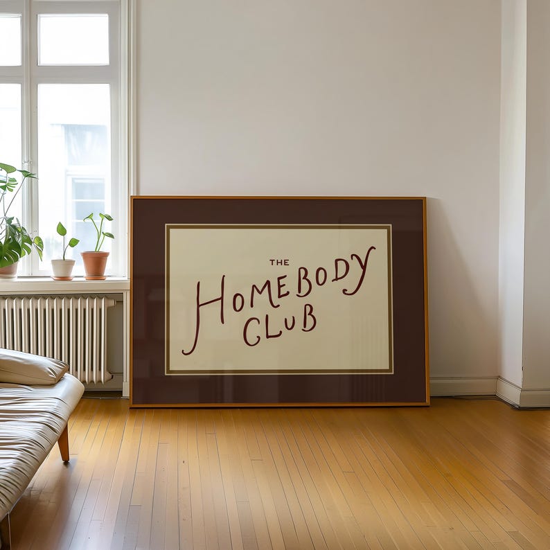 May include: A framed artwork with the text "The Homebody Club" in a handwritten style. The artwork is in a brown frame and is displayed on a wooden floor. The room has a window with plants and a radiator.