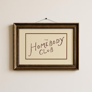May include: A framed print with the words "The Homebody Club" in a handwritten style. The text is in a deep red color against a cream-colored background. The frame is a dark, ornate brown.