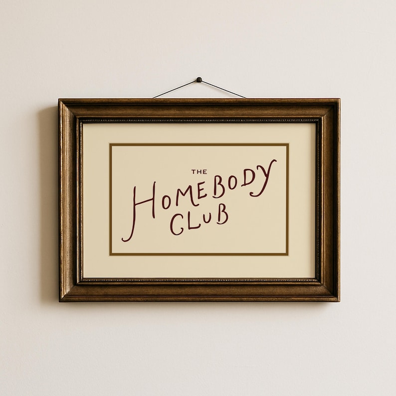 May include: A framed print with the words "The Homebody Club" in a handwritten style. The text is in a deep red color against a cream-colored background. The frame is a dark, ornate brown.