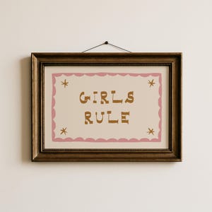 May include: A framed print with the words "GIRLS RULE" in brown letters on a cream background. The print has a pink scalloped border and star accents. The frame is a dark brown colour.