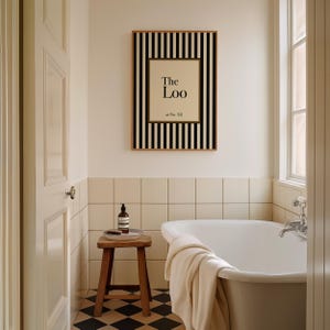 May include: A bathroom scene featuring a framed print with the text "The Loo" and a black and white striped border. A wooden stool holds a bottle and a small tray. A white bathtub and a window are also visible.