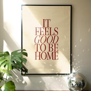 May include: A framed poster with the text "IT FEELS GOOD TO BE HOME" in burgundy lettering. The poster is set against a beige background and framed in black. A disco ball and a potted plant are on a wooden surface.