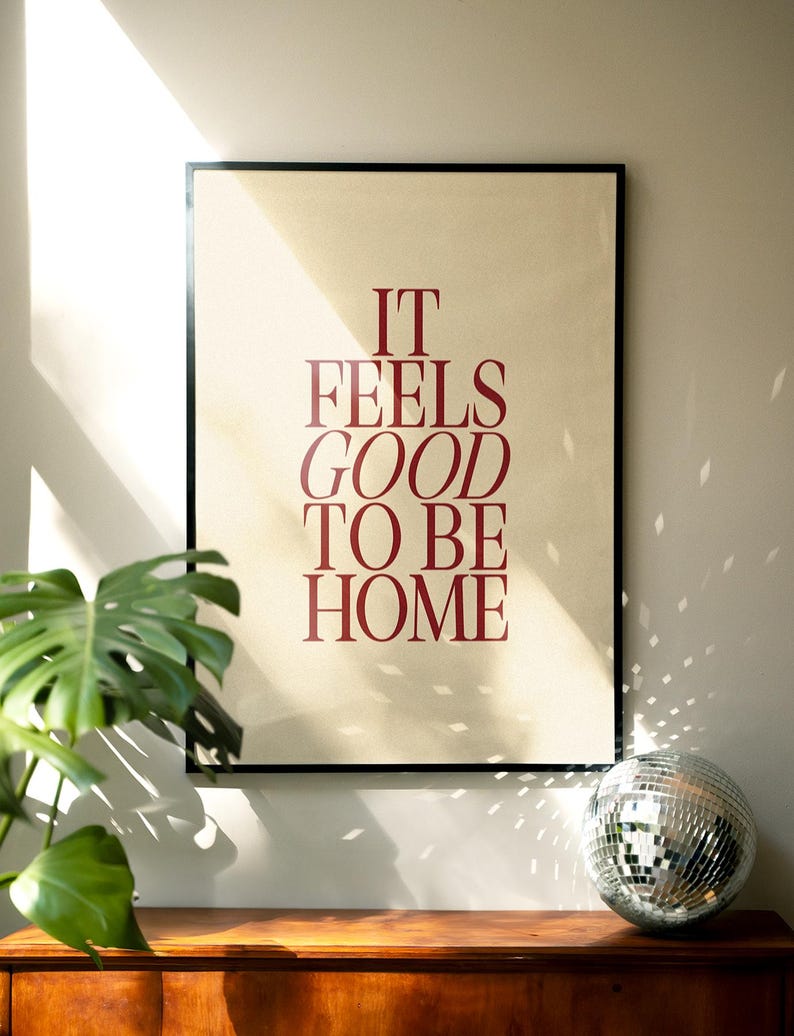 May include: A framed poster with the text "IT FEELS GOOD TO BE HOME" in burgundy lettering. The poster is set against a beige background and framed in black. A disco ball and a potted plant are on a wooden surface.