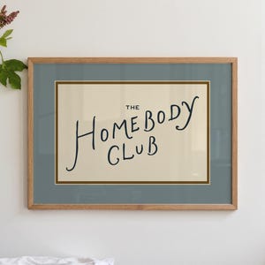 May include: A framed print with the words "THE Homebody CLUB" in black script on a cream background. The frame is light brown, and the matting is a muted blue-green. The print is a decorative item.