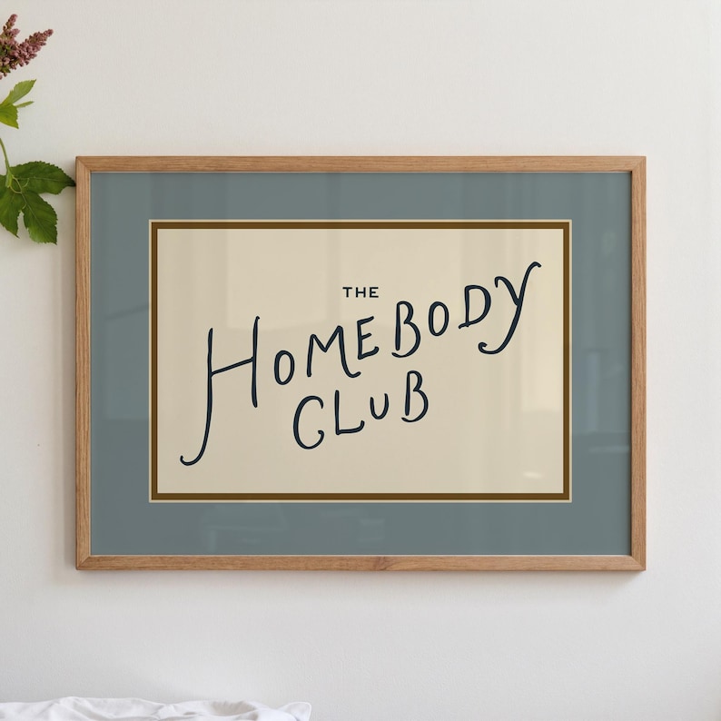 May include: A framed print with the words "THE Homebody CLUB" in black script on a cream background. The frame is light brown, and the matting is a muted blue-green. The print is a decorative item.