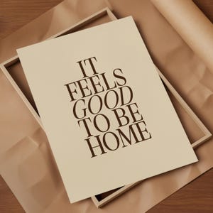 May include: A framed print with the text "IT FEELS GOOD TO BE HOME" in dark brown lettering. The print is a light beige color and is set inside a light wood frame. The background is a brown paper.