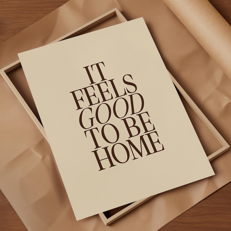 May include: A framed print with the text "IT FEELS GOOD TO BE HOME" in dark brown lettering. The print is a light beige color and is set inside a light wood frame. The background is a brown paper.
