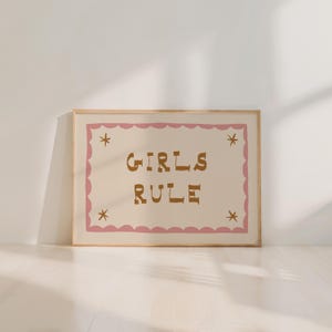 May include: A framed print with the words "GIRLS RULE" in brown letters on a cream background. The text is inside a pink scalloped border with star accents. The frame is a light wood color.