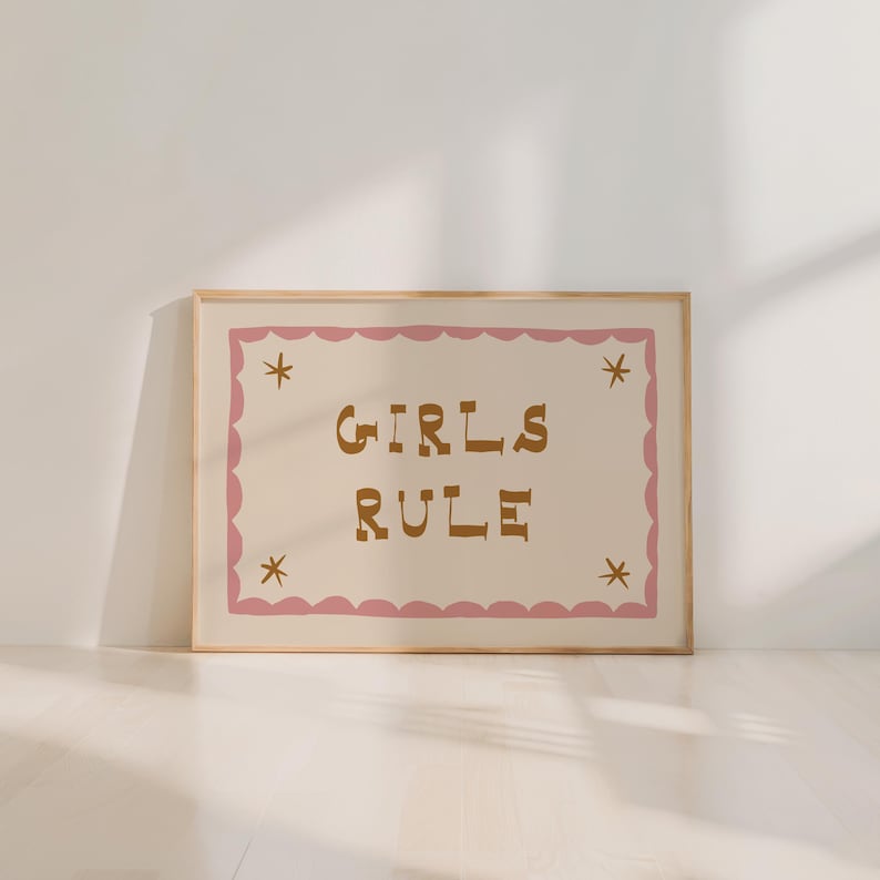 May include: A framed print with the words "GIRLS RULE" in brown letters on a cream background. The text is inside a pink scalloped border with star accents. The frame is a light wood color.