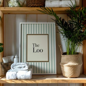 Sage green striped “The Loo” bathroom wall art styled on a wooden shelf with plants and towels