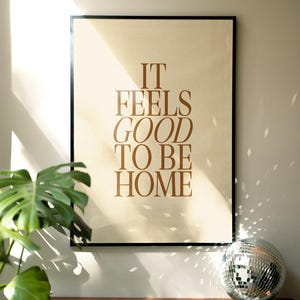 May include: Framed poster with the text "IT FEELS GOOD TO BE HOME" in brown lettering. The poster is displayed on a wooden surface, with a disco ball and a green plant in the foreground. The poster has a black frame.