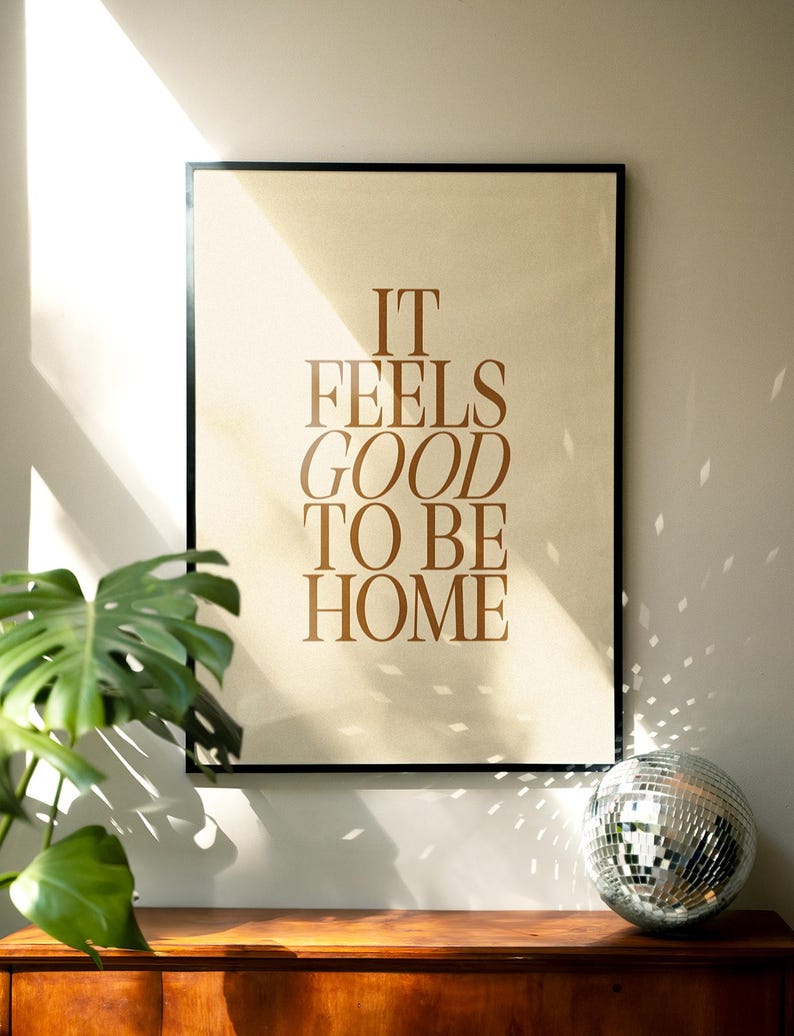 May include: Framed poster with the text "IT FEELS GOOD TO BE HOME" in brown lettering. The poster is displayed on a wooden surface, with a disco ball and a green plant in the foreground. The poster has a black frame.