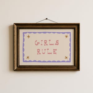 May include: A framed print with the words "GIRLS RULE" in pink letters. The print has a light beige background with a purple scalloped border and star accents. The frame is a dark brown color.