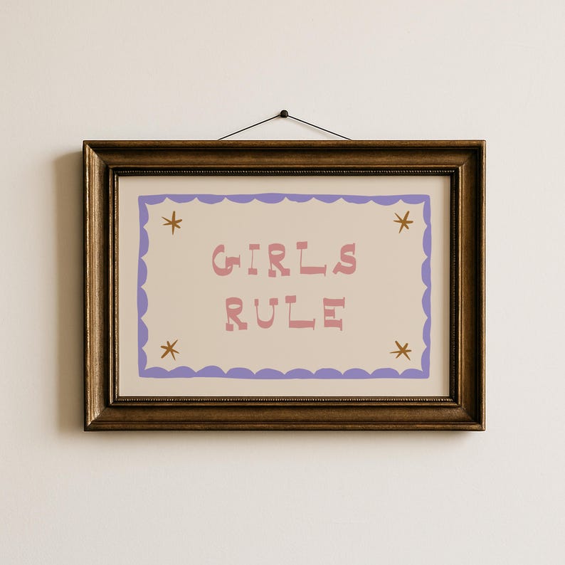 May include: A framed print with the words "GIRLS RULE" in pink letters. The print has a light beige background with a purple scalloped border and star accents. The frame is a dark brown color.
