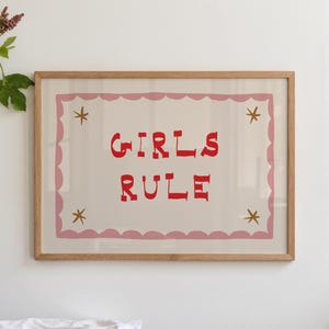 May include: A framed print with the words "GIRLS RULE" in red, centered on a cream background. The print is bordered by a pink scalloped edge and gold star accents, encased in a natural wood frame.