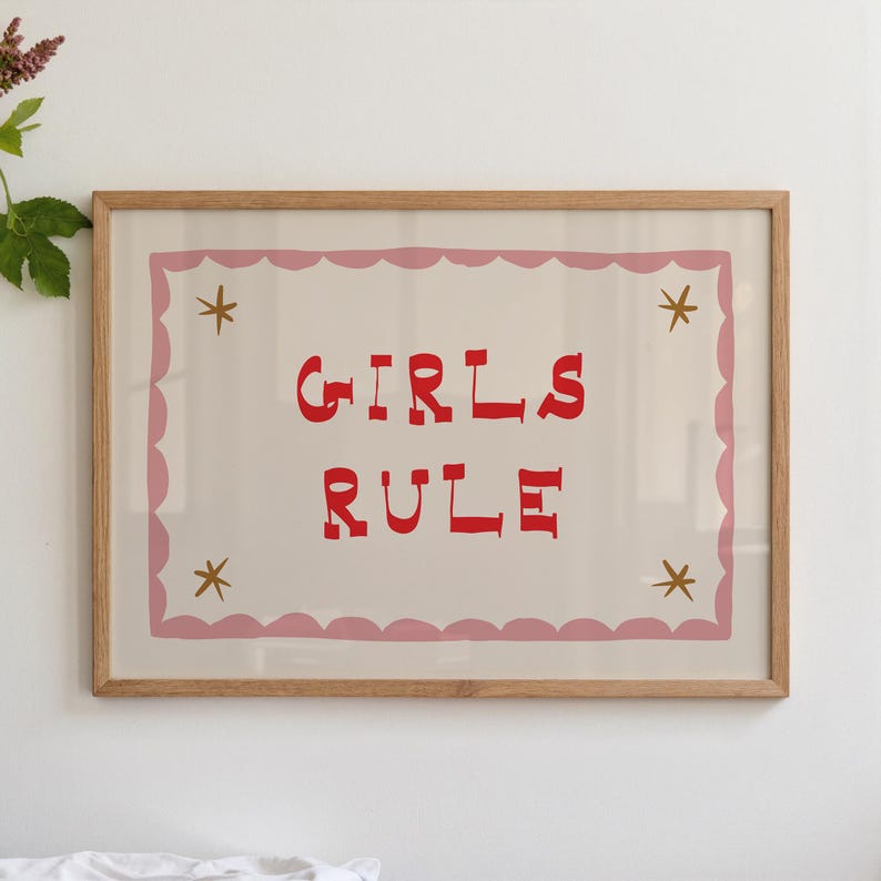 May include: A framed print with the words "GIRLS RULE" in red, centered on a cream background. The print is bordered by a pink scalloped edge and gold star accents, encased in a natural wood frame.