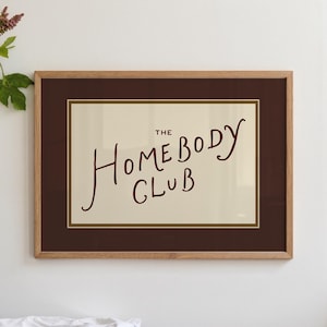 May include: A framed print with the words "THE HOMEBODY CLUB" in a handwritten style. The text is in a dark red color against a cream background, framed by a dark brown mat and a light brown wooden frame. The print is displayed on a white wall.