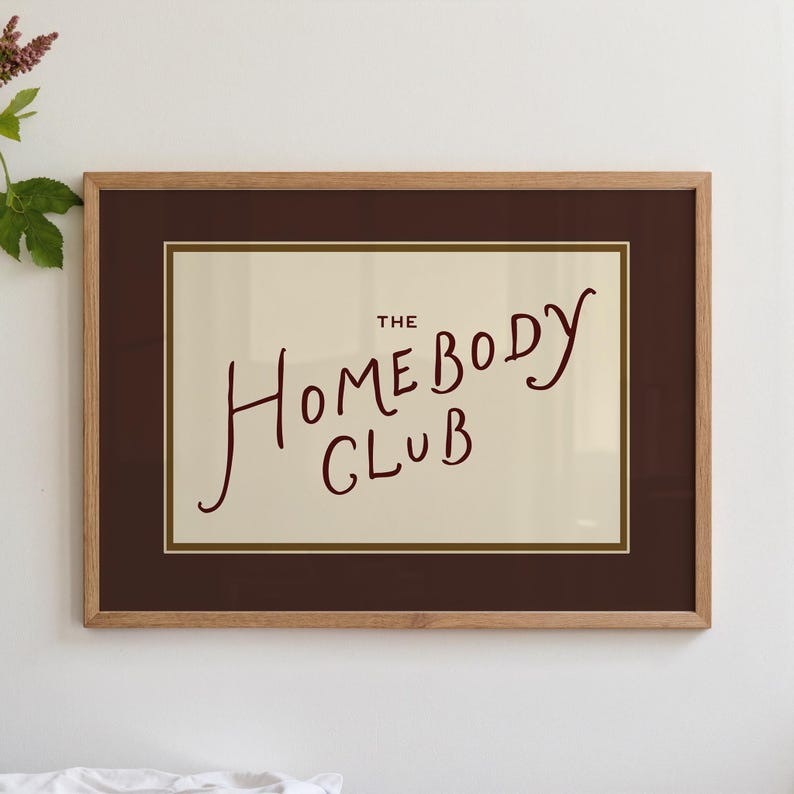 May include: A framed print with the words "THE HOMEBODY CLUB" in a handwritten style. The text is in a dark red color against a cream background, framed by a dark brown mat and a light brown wooden frame. The print is displayed on a white wall.