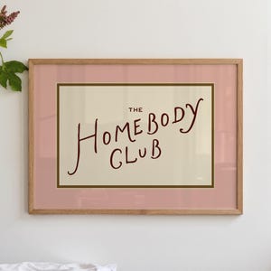 May include: A framed art print with the words "THE HOMEBODY CLUB" in a handwritten style. The print has a light pink border, a cream-colored background, and a wooden frame.