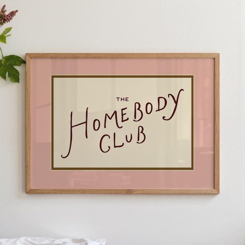 May include: A framed art print with the words "THE HOMEBODY CLUB" in a handwritten style. The print has a light pink border, a cream-colored background, and a wooden frame.