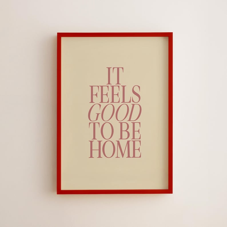 May include: A framed print with a red border. The print has a cream-colored background with the words "IT FEELS GOOD TO BE HOME" in a reddish-purple font. The artwork is displayed on a white wall.