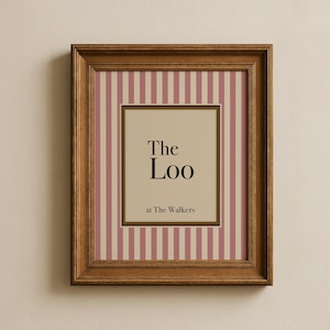 May include: A framed print with a brown wooden frame. The print has a cream background with pink vertical stripes. The center features a cream square with the words "The Loo" and "at The Walkers" in black text.