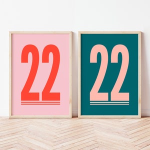 以下が含まれることがあります： 太字で大きく書かれた数字「22」が描かれた額装されたプリントが2つ。左側のプリントはピンクの背景に赤い数字、右側のプリントは緑がかった青の背景にピンクの数字が書かれている。