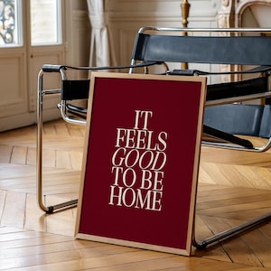 May include: A framed art print with the text "IT FEELS GOOD TO BE HOME" in white, set against a deep red background. The wooden frame is a light brown. The print is leaning against a chair on a hardwood floor.