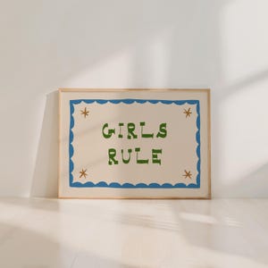 May include: A framed art print with the words "GIRLS RULE" in green, stylized lettering. The text is centered on a cream-colored background, framed by a blue scalloped border with star accents. The frame is a light wood color.