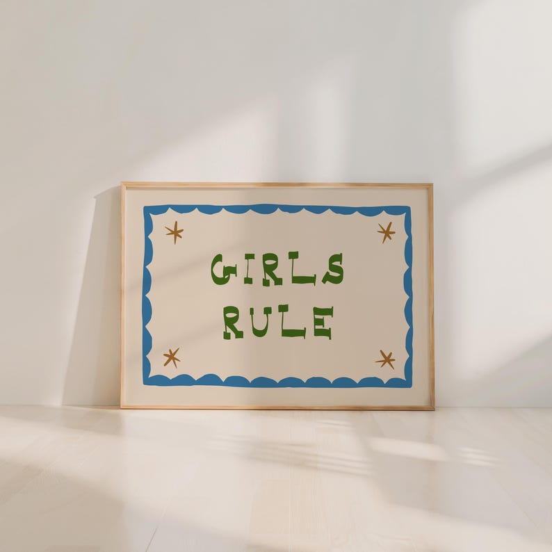 May include: A framed art print with the words "GIRLS RULE" in green, stylized lettering. The text is centered on a cream-colored background, framed by a blue scalloped border with star accents. The frame is a light wood color.