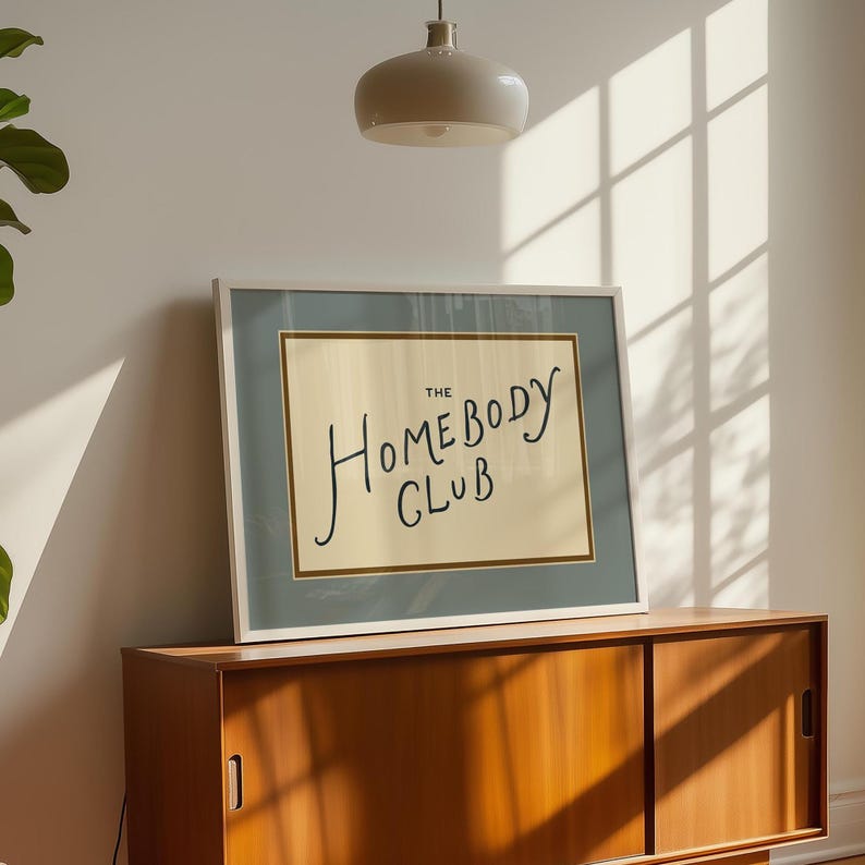 May include: A framed print with the text "The Homebody Club" in a handwritten style. The print has a light blue border and a cream-colored background. It is displayed on a wooden cabinet.