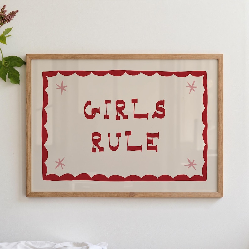 May include: A framed print with the words "GIRLS RULE" in red block letters. The text is centered on a cream-colored background, framed by a red scalloped border and four pink star accents. The frame is made of light-colored wood.