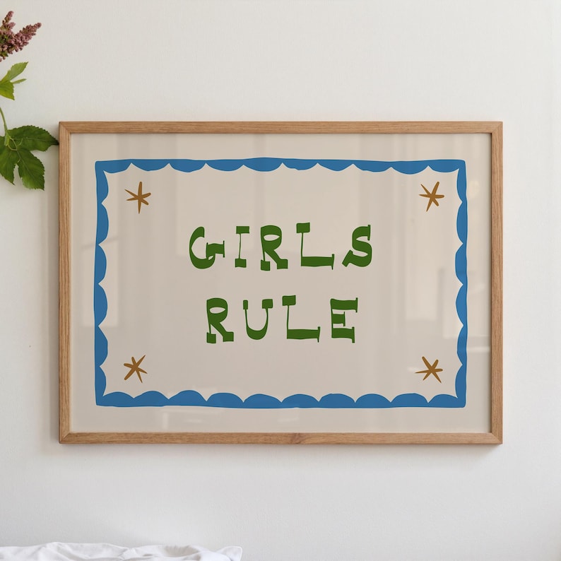 May include: A framed print with the words "GIRLS RULE" in green block letters. The text is centered on a cream-colored background, framed by a blue scalloped border and accented with small brown star shapes. The frame is made of light-colored wood.