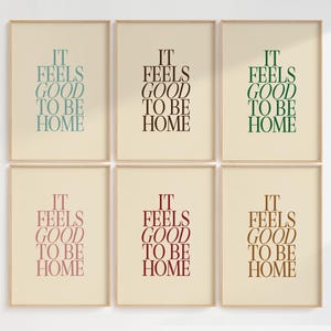 May include: Six framed art prints with the text "IT FEELS GOOD TO BE HOME" in various colors: teal, black, green, pink, burgundy, and brown. The text is centered on a cream-colored background, framed in a light wood.
