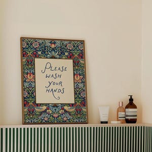 May include: A framed sign with the text "PLEASE WASH YOUR HANDS" in elegant script. The frame features a detailed floral and bird pattern in shades of blue, red, and green. The sign is displayed on a shelf with toiletries.
