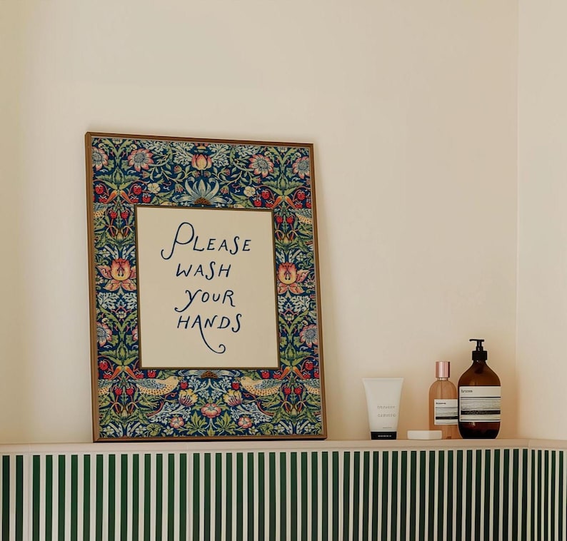 May include: A framed sign with the text "PLEASE WASH YOUR HANDS" in elegant script. The frame features a detailed floral and bird pattern in shades of blue, red, and green. The sign is displayed on a shelf with toiletries.