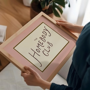May include: A framed print with the text "The Homebody Club" in a handwritten style. The print has a light pink border and a cream-colored background. The frame is made of light-colored wood.