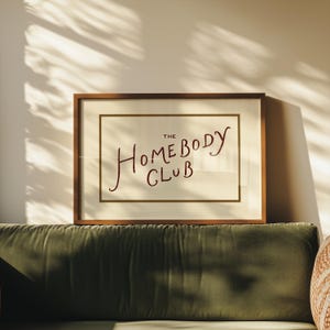 May include: A framed print with the text "The Homebody Club" in a handwritten style. The artwork is in a brown wooden frame with a cream-colored background. The print is displayed above a green velvet sofa.