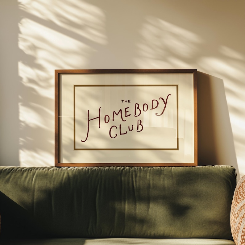 May include: A framed print with the text "The Homebody Club" in a handwritten style. The artwork is in a brown wooden frame with a cream-colored background. The print is displayed above a green velvet sofa.