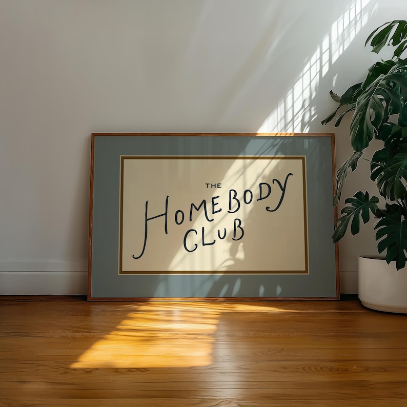 May include: A framed art print with the words "THE HOMEBODY CLUB" in black script. The print has a light beige background, a light blue border, and a wooden frame. The artwork is displayed on a wooden floor.
