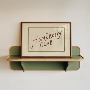 May include: A framed print with the words "The Homebody Club" in a handwritten style, displayed on a two-tiered green and wood-toned shelf. The shelf is mounted on a white wall, creating a minimalist aesthetic.