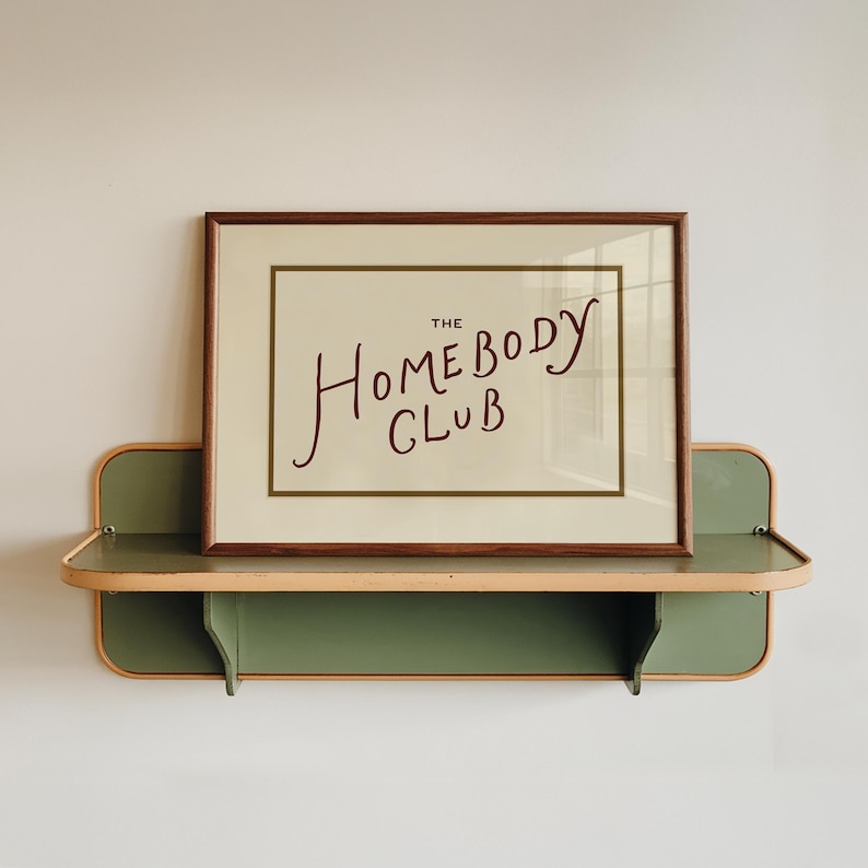 May include: A framed print with the words "The Homebody Club" in a handwritten style, displayed on a two-tiered green and wood-toned shelf. The shelf is mounted on a white wall, creating a minimalist aesthetic.