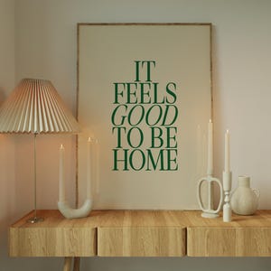 May include: A wooden console table with a framed print that reads "IT FEELS GOOD TO BE HOME" in green lettering. A pleated lampshade, candles, and decorative vases complete the home decor scene.