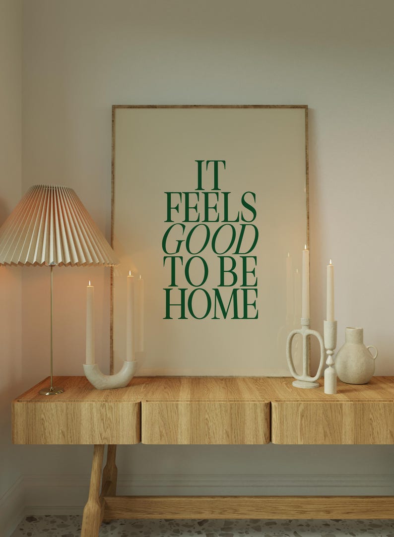 May include: A wooden console table with a framed print that reads "IT FEELS GOOD TO BE HOME" in green lettering. A pleated lampshade, candles, and decorative vases complete the home decor scene.